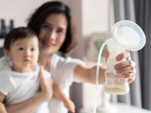 Happy Asian mom holding her breast pump milk and cuddling her baby to illustrate possible challenges when breastfeeding
